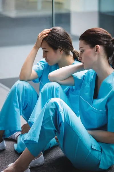 Two female healthcare workers in scrubs sitting on the floor, resting during a busy shift.